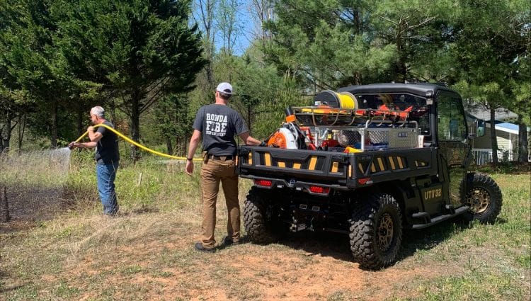 Two People Standing Next To An Off-Road Vehicle. One Person Is Holding A Hose And Spraying The Ground.